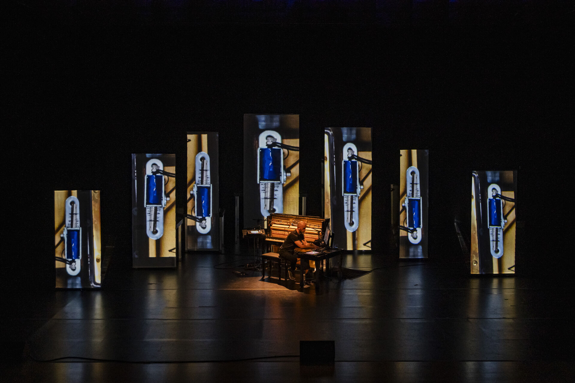 Expanded Piano II stage view: an illuminated piano centred on stage, framed by an arc of seven tall standing panels each showing a vertical blue-toned video projection. Photo by Andreas Simopoulos.
